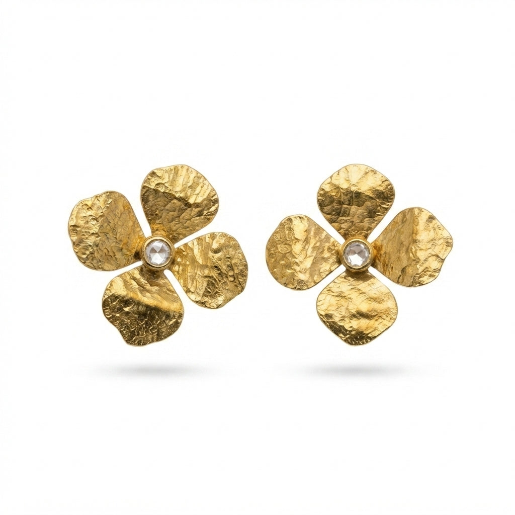 A pair of textured gold flower-shaped earrings with small clear center stones, displayed on a white marble pedestal with a gold rim against a softly blurred beige background.