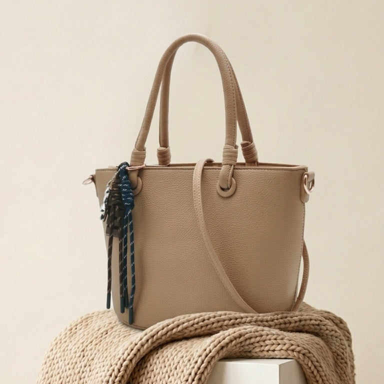 A taupe leather tote bag with dual top handles and a dark blue braided tassel charm, styled on a white pedestal draped with a chunky beige knit blanket against a warm beige background.