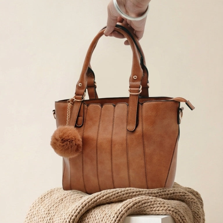A brown leather tote bag with a fluffy pom-pom charm, held by a hand and draped over a chunky knit beige throw blanket on a white pedestal against a warm beige background.
