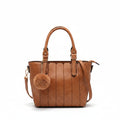 A tan leather tote bag with a fluffy pom-pom charm, held by a hand and draped over a chunky knit beige throw blanket on a white pedestal against a warm beige background.