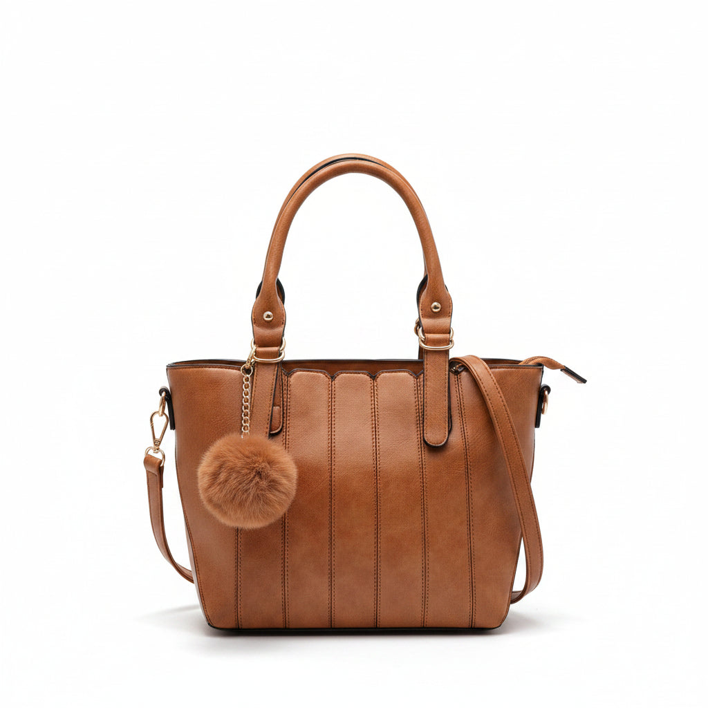 A tan leather tote bag with a fluffy pom-pom charm, held by a hand and draped over a chunky knit beige throw blanket on a white pedestal against a warm beige background.