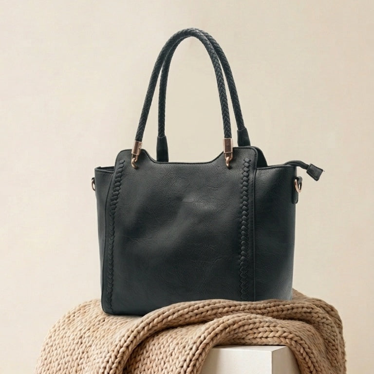 A structured black leather tote bag with double top handles and brass-tone hardware, displayed on a white pedestal draped with a chunky beige knit throw blanket against a warm beige background.