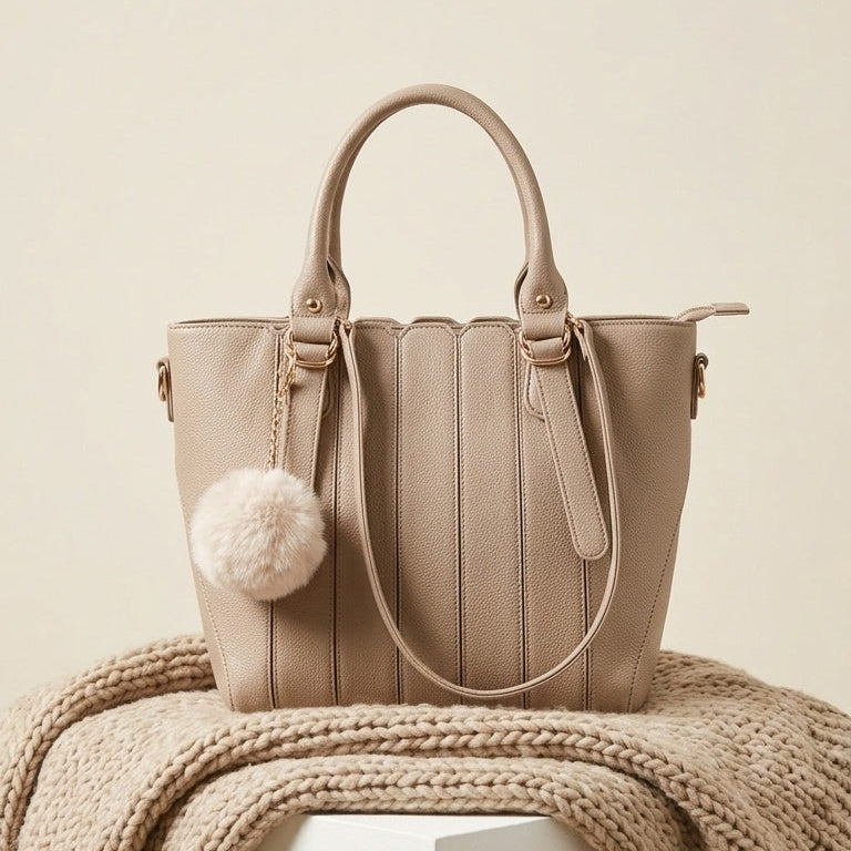 A structured beige tote bag with dual top handles, a hanging fluffy pom-pom charm, and a shoulder strap, displayed on a white pedestal draped with a chunky knit beige throw against a warm cream background.