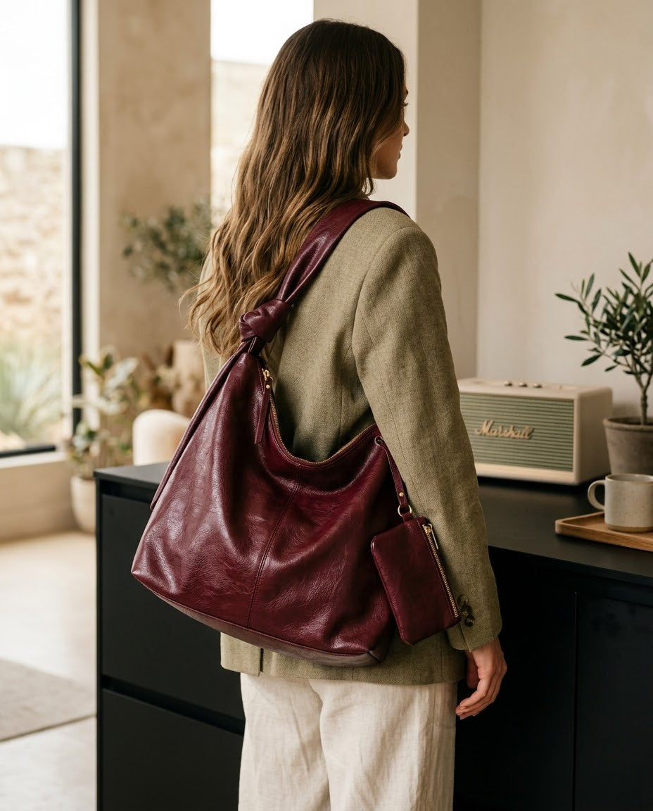 A woman wearing a sage green cardigan and cream pants is shown from behind, carrying a large burgundy leather hobo shoulder bag with a matching zippered pouch. The setting is a modern, sunlit room with a black sideboard, a vintage Marshall speaker, and potted plants.