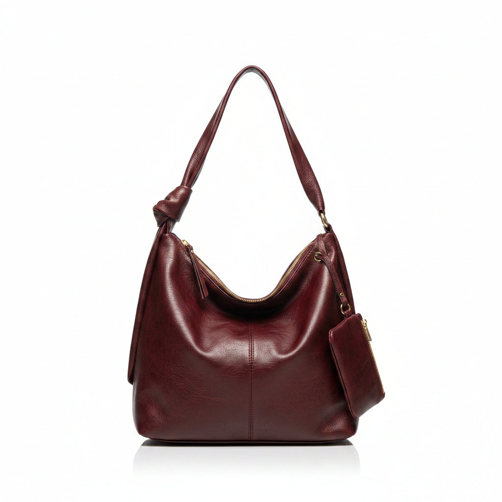 A woman wearing a sage green cardigan and cream pants is shown from behind, carrying a large burgundy leather hobo shoulder bag with a matching zippered pouch. The setting is a modern, sunlit room with a black sideboard, a vintage Marshall speaker, and potted plants.