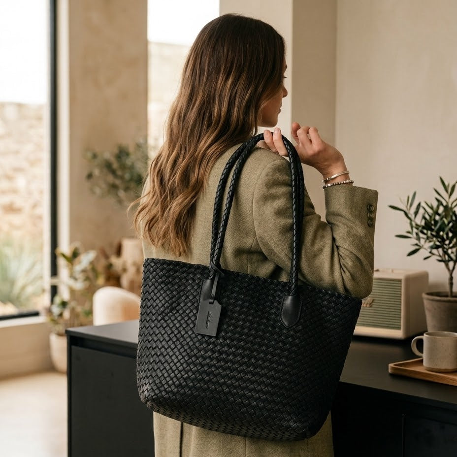 A woman viewed from behind wearing a black woven tote bag with braided shoulder straps, standing indoors near a window and counter.