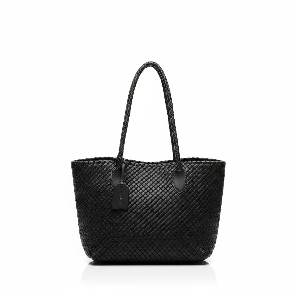A woman viewed from behind wearing a black woven tote bag with braided shoulder straps, standing indoors near a window and counter.