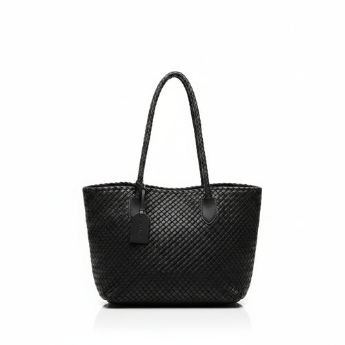 A woman viewed from behind wearing a black woven tote bag with braided shoulder straps, standing indoors near a window and counter.