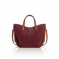 A styled flat-lay of a burgundy suede tote bag with brown leather handles, surrounded by matching beige and brown accessories on a warm beige background. The bag is shown with a phone, sunglasses, earbuds, bottles, a wallet, and a keychain.