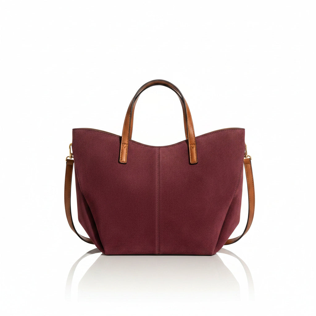 A styled flat-lay of a burgundy suede tote bag with brown leather handles, surrounded by matching beige and brown accessories on a warm beige background. The bag is shown with a phone, sunglasses, earbuds, bottles, a wallet, and a keychain.