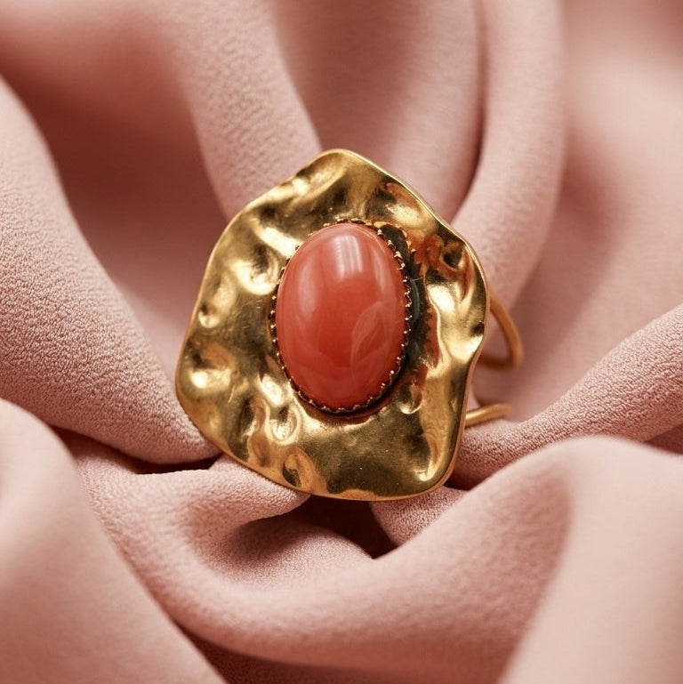 A gold ring with a wavy, organic-shaped setting and a smooth, oval coral-pink stone, displayed on draped dusty pink fabric.