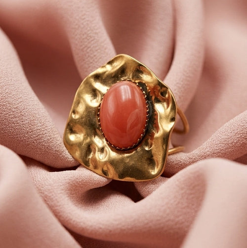 A gold ring with a wavy, organic-shaped setting and a smooth, oval coral-pink stone, displayed on draped dusty pink fabric.