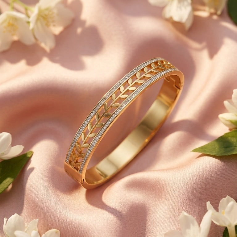 A gold bangle bracelet with a textured chevron pattern and a row of small clear stones, styled on a pink satin fabric with white flowers and green leaves.