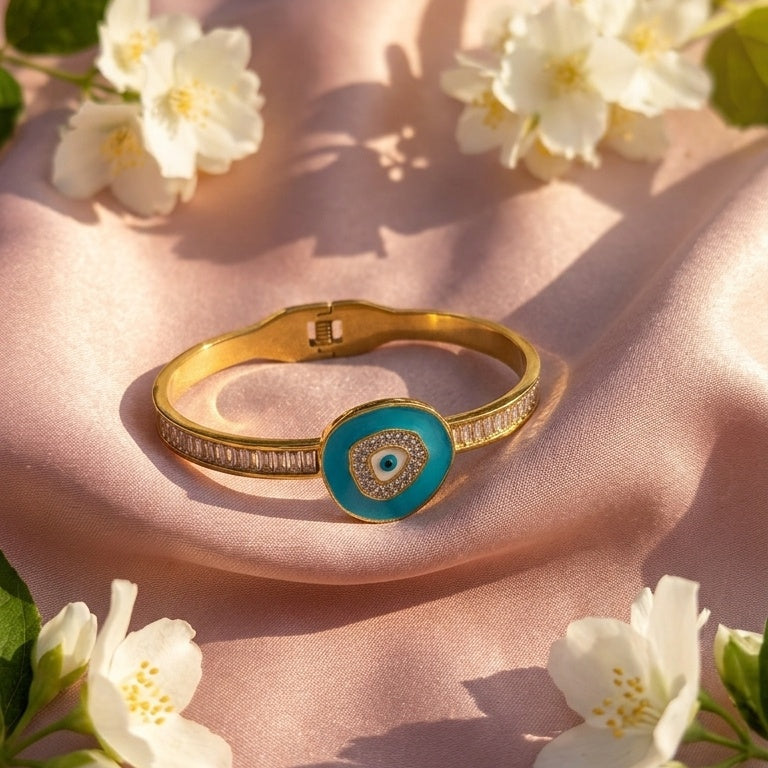 A gold bangle bracelet with a turquoise blue circular evil eye pendant, styled on draped pink satin fabric with white flowers.
