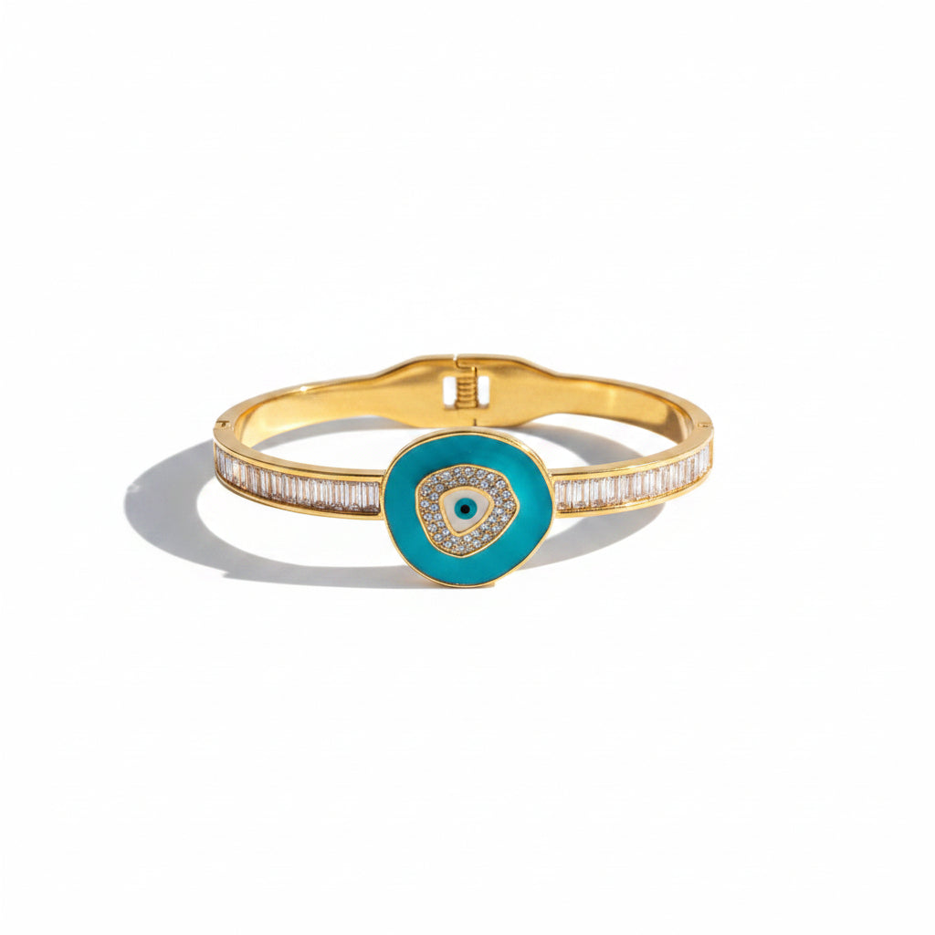 A gold bangle bracelet with a turquoise blue circular evil eye pendant, styled on draped pink satin fabric with white flowers.