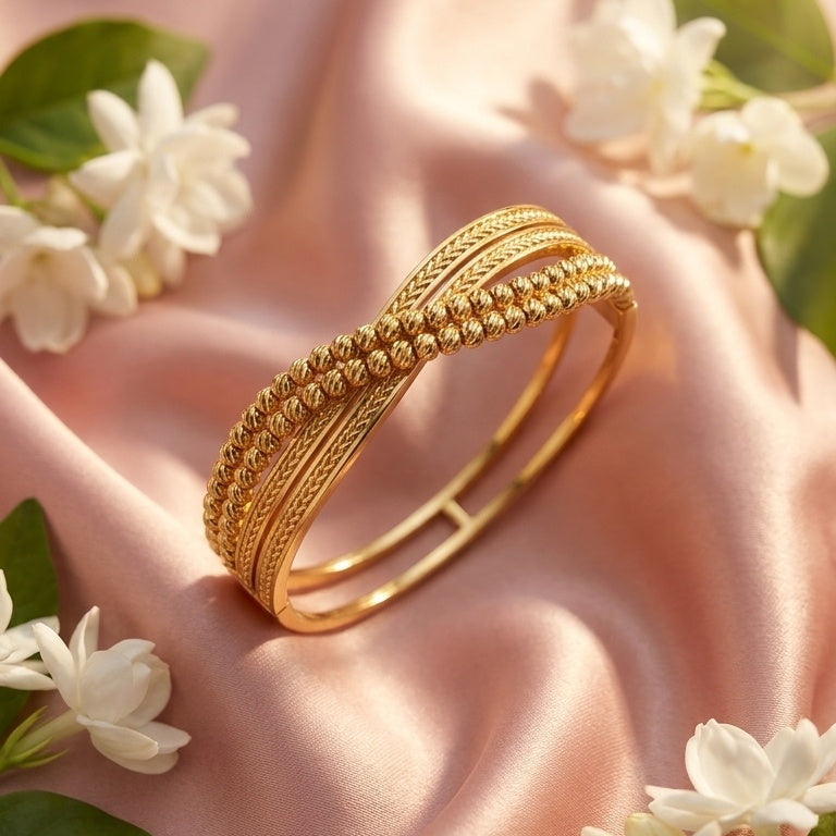A gold-toned multi-band bangle bracelet with textured and woven details, displayed on draped pink satin fabric with white flowers and green leaves around it.