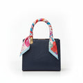 A navy blue leather handbag with a colorful floral silk scarf tied to its top handle, shown against a plain white background.