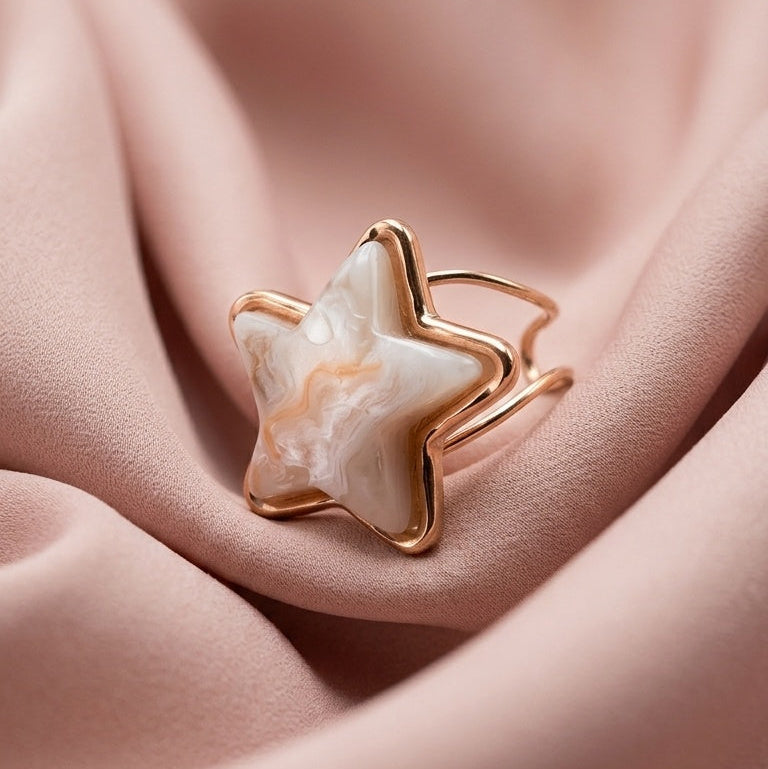 A gold star-shaped ring with a white and pink marbled stone, resting on draped dusty pink satin fabric.