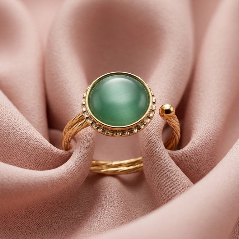 A gold ring with a round green cabochon gemstone and small clear accent stones, displayed on soft pink satin fabric.