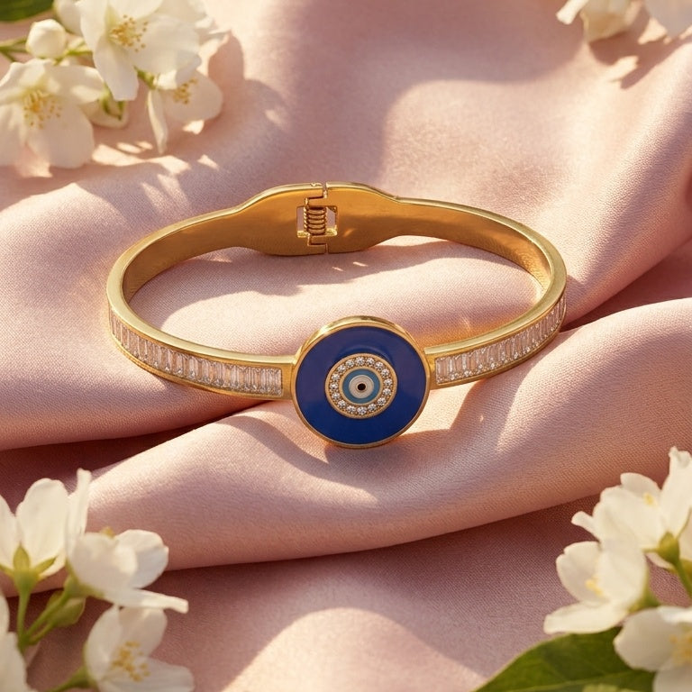 A gold bangle bracelet with a blue and white evil eye emblem and a row of small clear stones, styled on pink satin fabric with white flowers.