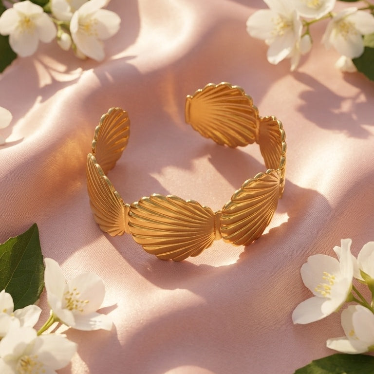 A gold-toned open cuff bracelet with ribbed shell-like segments, styled on a soft pink satin fabric with white flowers and green leaves.