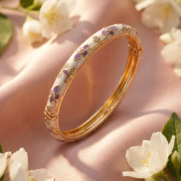A gold bangle bracelet with purple and white floral enamel detailing, styled on a draped pink fabric with white flowers.