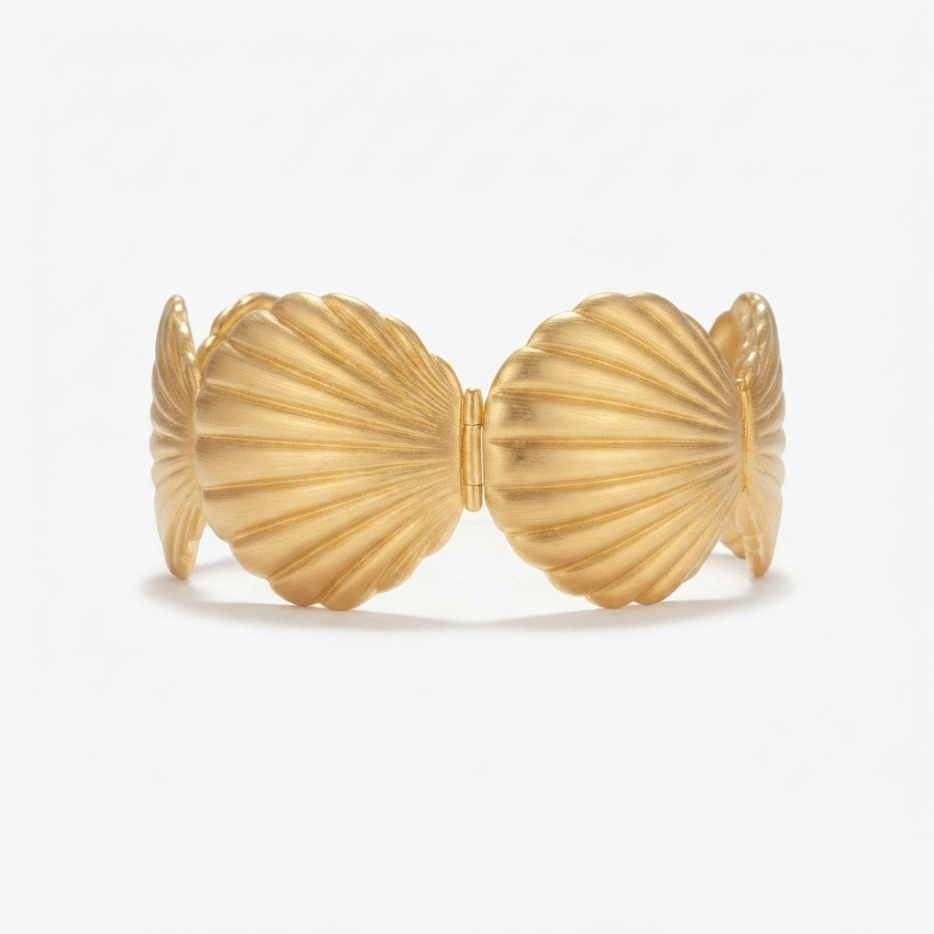 A gold-toned open cuff bracelet with ribbed shell-like segments, styled on a soft pink satin fabric with white flowers and green leaves.