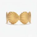 A gold-toned open cuff bracelet with ribbed shell-like segments, styled on a soft pink satin fabric with white flowers and green leaves.