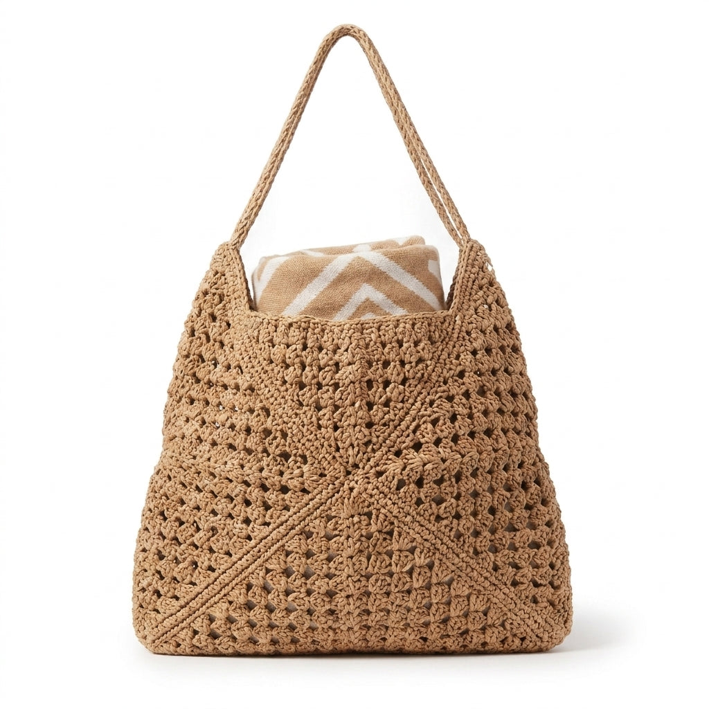 A tan crochet shoulder bag with a woven rope handle, shown on a clean white background with a folded patterned blanket tucked inside.