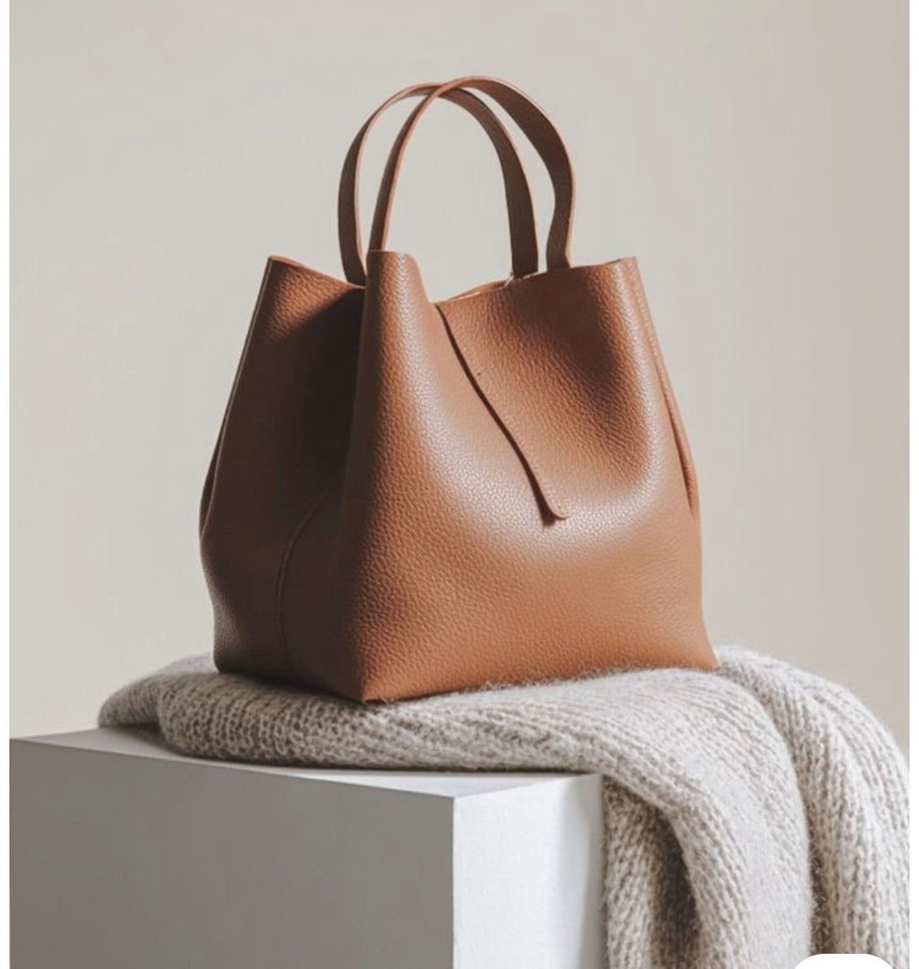 A medium brown pebbled leather tote bag with double top handles, resting on a chunky cream knit throw blanket atop a white display block against a warm beige background.