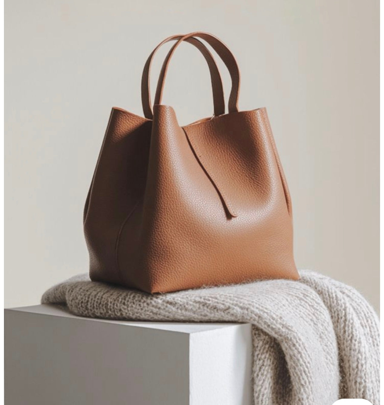 A medium brown pebbled leather tote bag with double top handles, resting on a chunky cream knit throw blanket atop a white display block against a warm beige background.