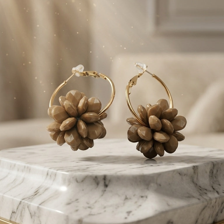 A pair of gold hoop earrings with large, textured brown flower-like dangle charms, displayed on a white and gray marble tray with a gold rim against a softly blurred beige background.