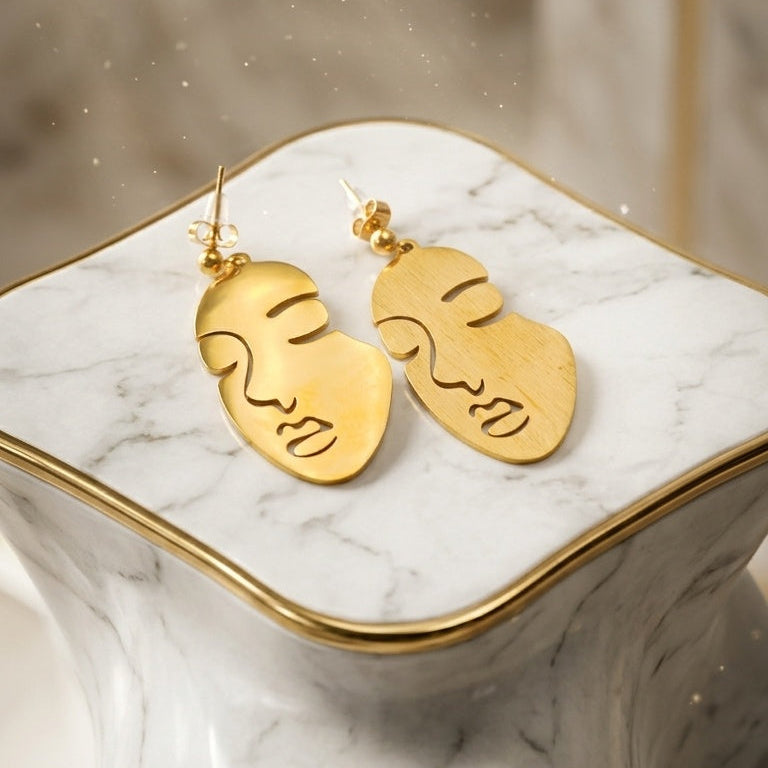 A pair of gold face-shaped dangle earrings with engraved facial features, displayed on a white and gray marble side table with gold trim.