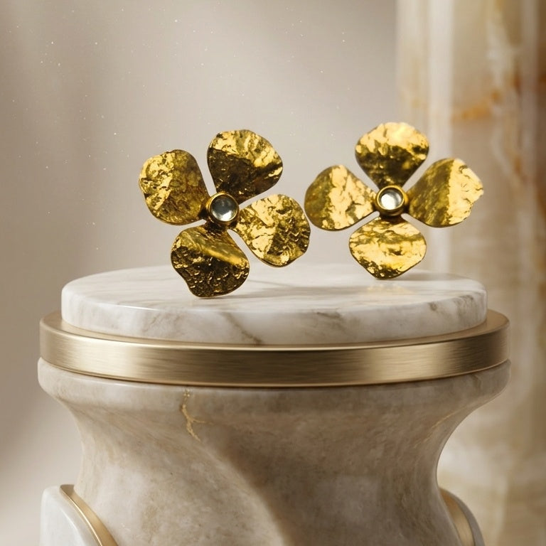 A pair of textured gold flower-shaped earrings with small clear center stones, displayed on a white marble pedestal with a gold rim against a softly blurred beige background.